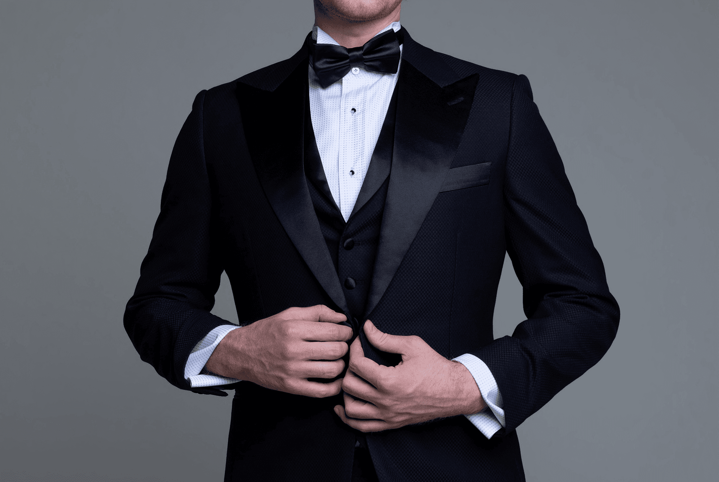 Man in a black tuxedo with satin lapels and bow tie fastening his jacket against a gray studio background.
