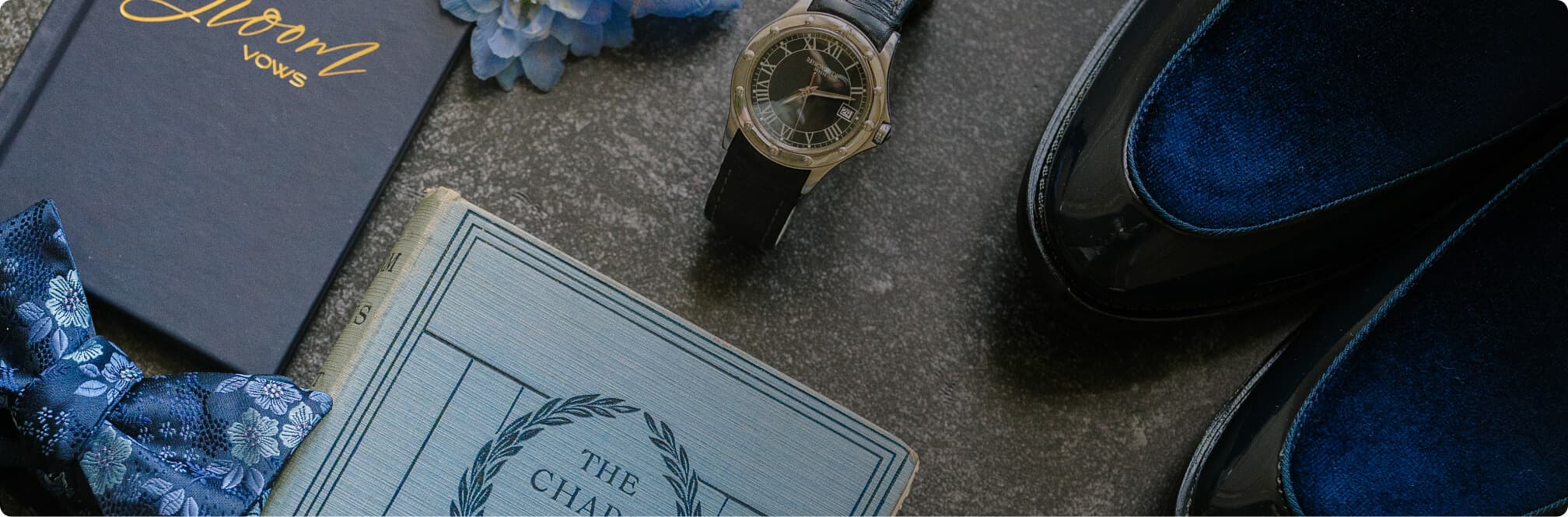 Flat lay of blue velvet shoes, a watch, a bow tie, and wedding accessories.
