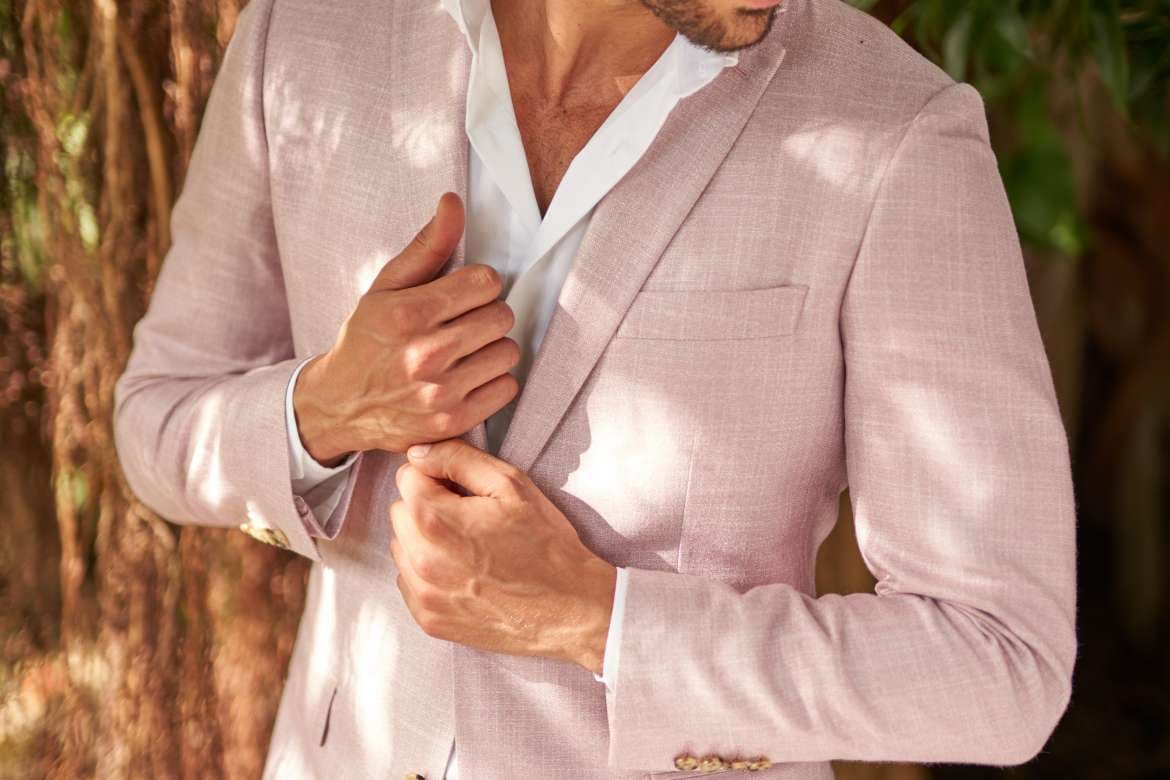 Close-up of a man buttoning a light pink blazer over a white shirt outdoors.
