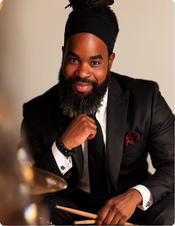 Bearded man in a black suit holding drumsticks beside a cymbal.