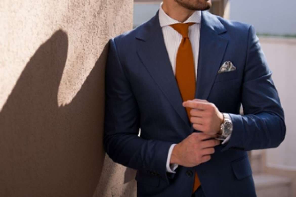 Close-up of a man in a blue suit adjusting his cuff with an orange tie and pocket square.