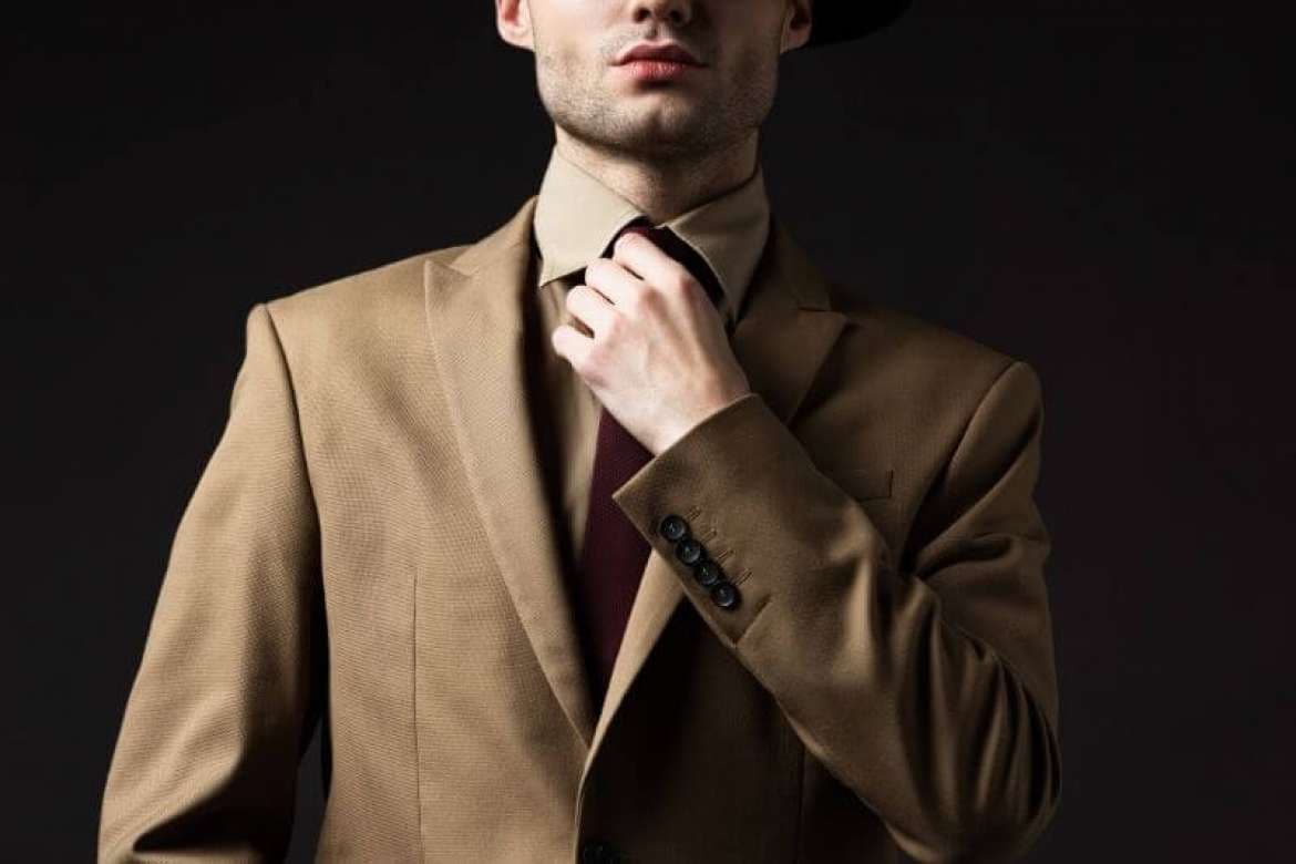 Man in a tan suit adjusting his tie against a dark background.