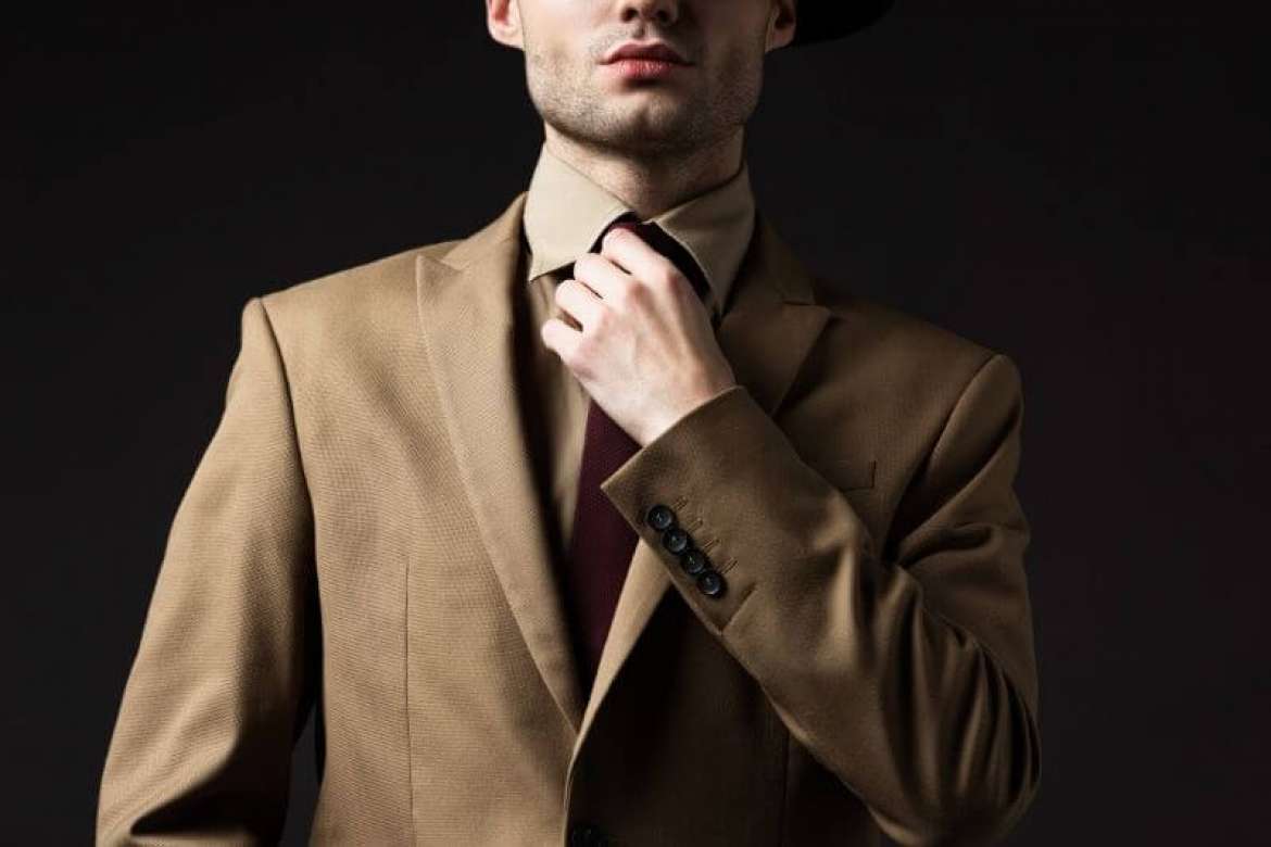 Man in a tan suit adjusting his tie against a dark background.