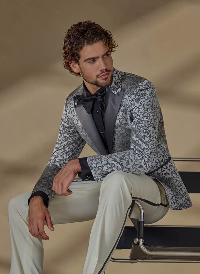 Man seated in a silver patterned tuxedo jacket with black bow tie.
