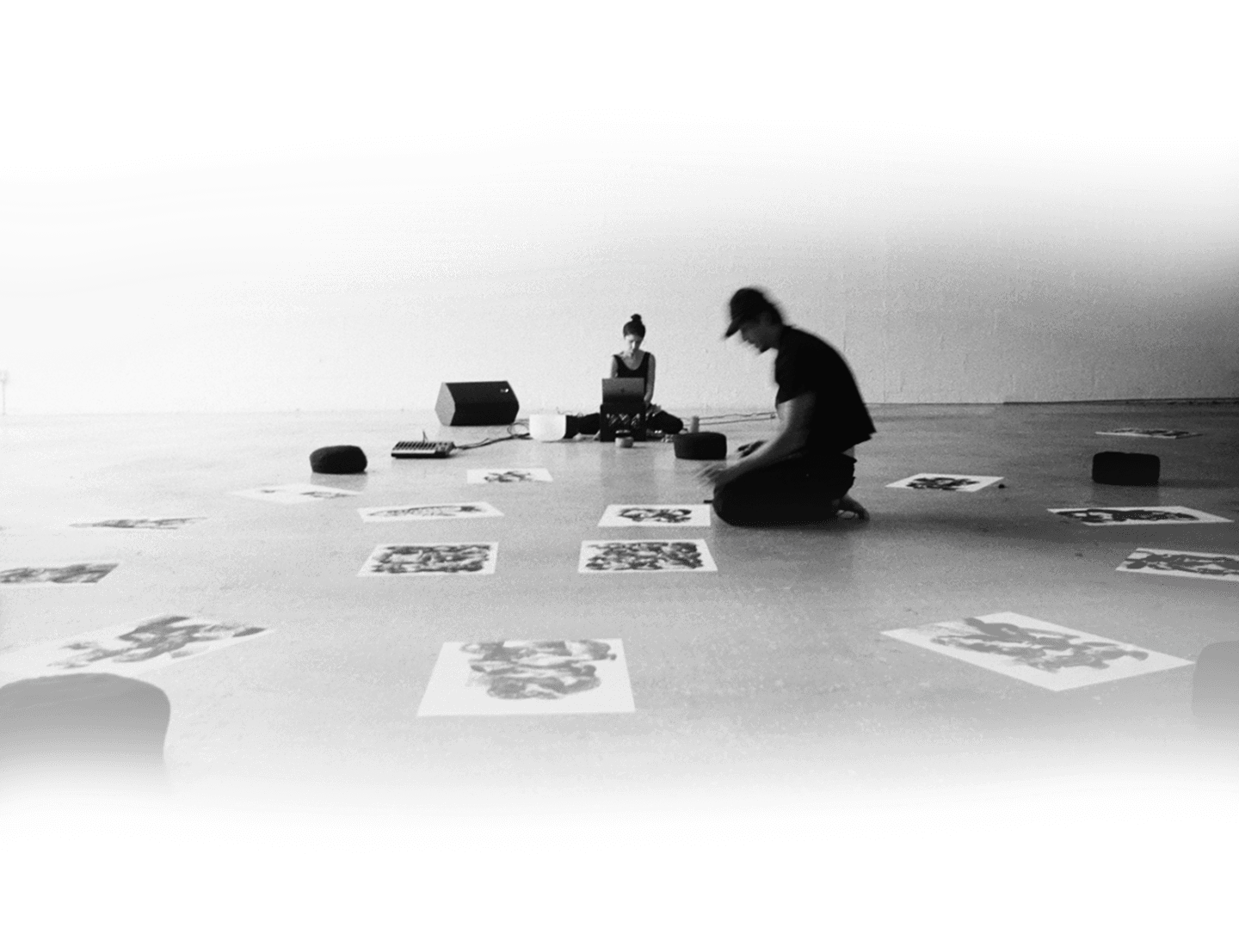 Black-and-white studio scene with an artist seated among printed artworks.