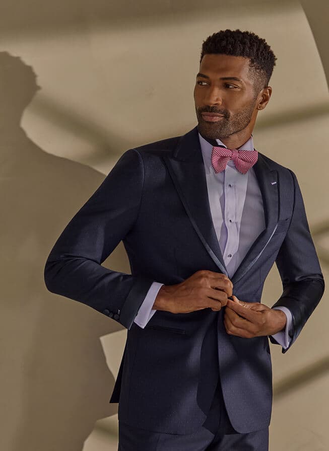 Man in a navy suit fastening his jacket with a pink bow tie.