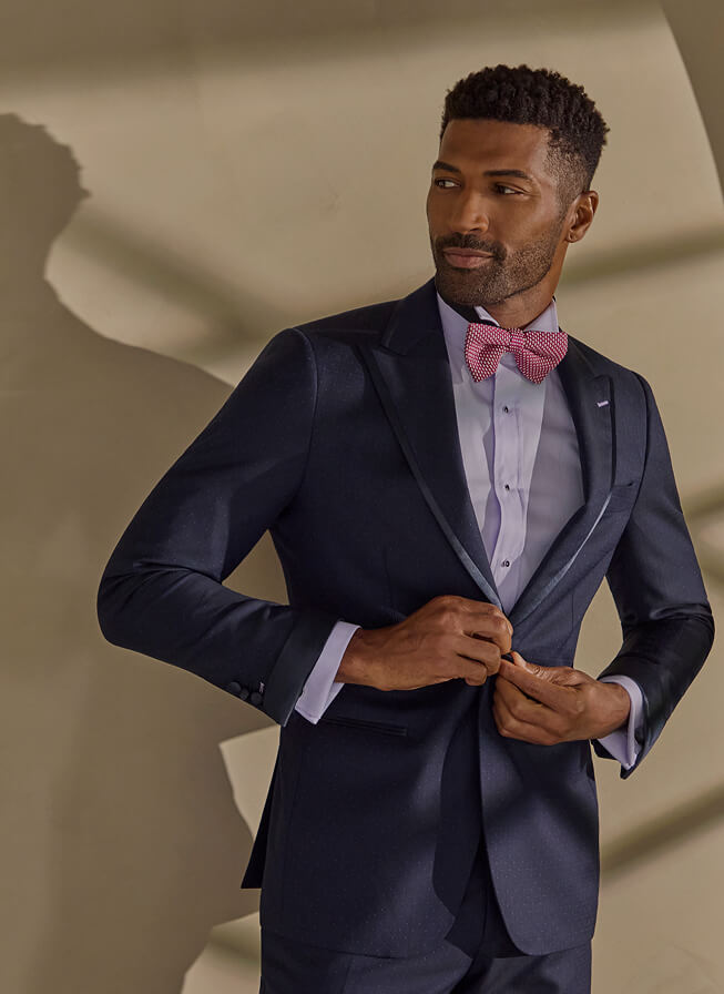 Man in a navy suit fastening his jacket with a pink bow tie.