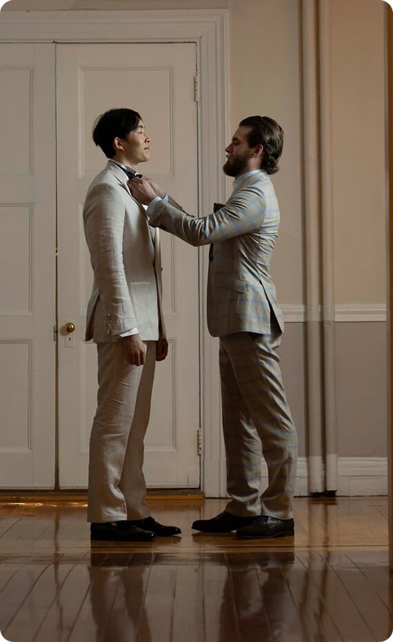 Man adjusting another man’s bow tie in a wedding suit fitting.