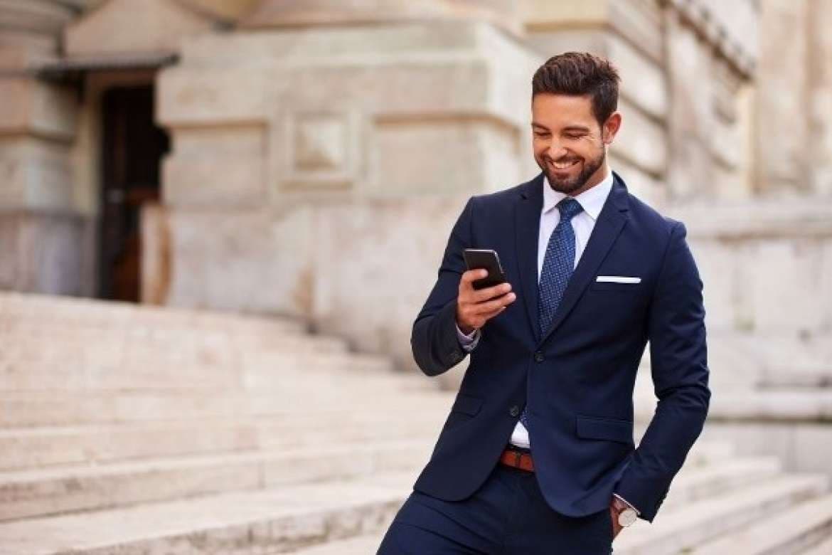 Smiling man in a navy suit checking his phone on outdoor stone steps.